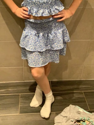 Blommig volangsätt i ljusblått  - Supersött sätt  i ljusblått med vita och blå blommor. Kjolen har två lager volanger och smock i midjan för en bekväm passform. Perfekt för dig som gillar en feminin och lekfull stil. Tunn och luftig, passar perfekt till varma dagar. Storlek 134/140