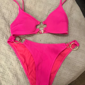 Rosa ribbad bikini New Yorker  - Snygg rosa bikini  med ribbad struktur. Toppen har triangelformade kupor och smala axelband, samt guldfärgade ringar och en stjärndetalj framtill. Underdelen har högt skuren midja och matchande guldringar på sidorna. Perfekt för stranden eller poolen!