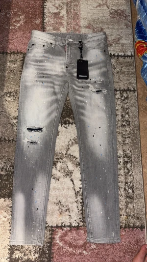 Grå Dsquared2 jeans med slitningar - Säljer ett par grå jeans från Dsquared2 med coola slitningar och färgstänk i vitt och blått. Modellen har smal passform, klassisk femficksdesign och är tillverkad i stretchig bomull. Perfekt för dig som gillar streetstyle och vill sticka ut.