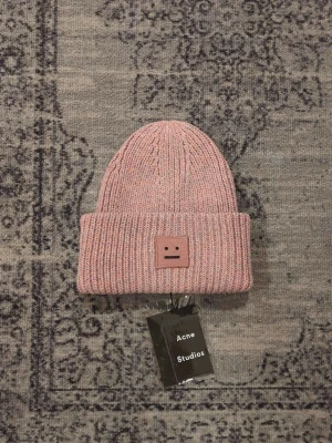 Rosa stickad mössa Acne Studios - Säljer en rosa stickad mössa från Acne Studios med det ikoniska ansiktsloggan framtill. Mössan är ribbstickad och gjord i 100% ull, vilket gör den både varm och snygg. Perfekt accessoar för att lyfta din outfit under kyliga dagar.