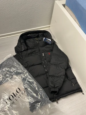 Svart dunjacka från Polo Ralph Lauren - Säljer en svart dunjacka från Polo Ralph Lauren med klassisk röd logga på bröstet. Jackan har hög krage, flera dragkedjeförsedda fickor och vattenavvisande material. Perfekt för kalla dagar och riktigt snygg passform. Det finns också en ficka på insidan av jackan. Överiga frågor eller om ni vill få mer bilder kontakta gärna mig ;) (Storlek S)