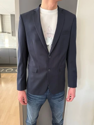 Mörkblå kavaj från Dressmann - Säljer en mörkblå kavaj från Dressmann i slim fit-modell. Kavajen har två knappar framtill, klassiska fickor med lock och en bröstficka. Insidan är fodrad med ljusblått tyg och röda detaljer. Perfekt för dig som vill ha en stilren och modern look.