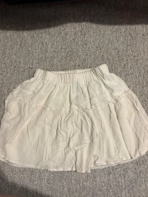 Dam Vit volangkjol - Supersöt kort vit kjol med volangdetaljer och bred resår i midjan i storlek XS. Kjolen har en luftig och flowy känsla, perfekt för varma dagar. Materialet känns lätt och mjukt mot huden. Kjolen är vit men ser lite gul ut pågrund av ljuset. 