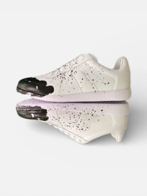 Maison Margiela sneakers med svart färgstänk - Unika vita sneakers från Maison Margiela i skinn med svart färgstänk och drip-detalj över tån. Skorna har klassisk snörning, rund tå och platt sula. Snygg kontrast mellan det vita och svarta, och boxen medföljer. Perfekt för dig som gillar statement-plagg.