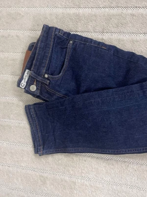 Mörkblå jeansbyxor från Circular - Säljer ett par mörkblå jeansbyxor från Circular med klassisk femficksdesign och kontrastsömmar. Byxorna har rak passform och normal midja. Materialet är tåligt jeans och de har en snygg läderpatch bak i midjan. Perfekta till din vardagsstil!