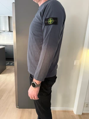 Grå långärmad tröja Stone Island - Säljer en grå långärmad tröja från Stone Island med klassisk patch på ärmen. Tröjan har knappar vid halsen och är tillverkad i ett strukturerat bomullstyg. Perfekt för dig som gillar stilrena och avslappnade plagg.