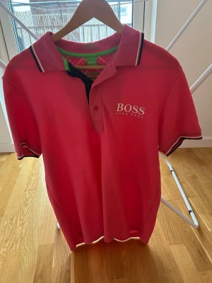 Rosa pikétröja från Hugo Boss M - Säljer en snygg rosa pikétröja från Hugo Boss med vit logga på bröstet. Tröjan har klassisk krage med kontrastfärgade ränder och knappar framtill. Insidan av kragen har coolt rutmönster och gröna detaljer. Perfekt för dig som vill ha en sportig och stilren look.