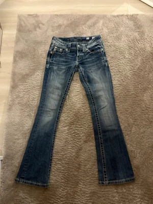 Miss Me bootcut jeans med bling - Säljer ett par blå Miss Me jeans med bootcut och snygga slitningar. Jeansen har låg midja, silverfärgade detaljer och blingade bakfickor med paljetter och nitar. Klassisk denim med coola detaljer och markerade sömmar. Perfekta för dig som gillar statement-jeans.