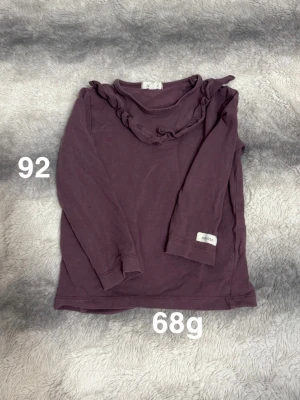 Lila långärmad topp med volang - Söt långärmad topp från Newbie i en mörklila nyans. Toppen har en rund halsringning dekorerad med volangdetaljer som ger en gullig touch. Perfekt för dig som gillar stilrena men ändå lekfulla plagg.🌸 Fint använt skick. Storlek 92