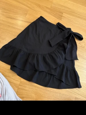 Svart omlottkjol med volang - Supersnygg svart omlottkjol med bred knytning i midjan och dubbla volanglager. Kjolen har en feminin och flowy look som passar perfekt till sneakers eller boots. Materialet känns  lätt, och modellen är normal i passformen.