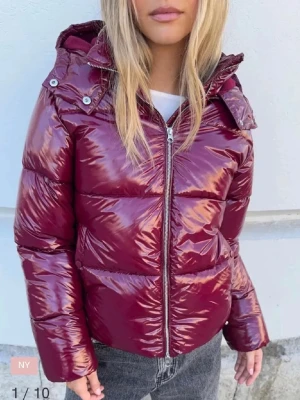 Nelly vinröd pufferjacka  - Säljer en glansig vinröd pufferjacka sopm har huva och dragkedja framtill. Jackan är vadderad och har en kortare, boxig passform som ger en cool look. Perfekt för kalla dagar när du vill sticka ut lite extra.