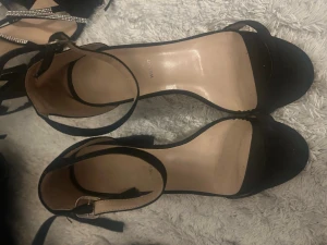 Svarta klackade sandaler med ankelrem - Säljer ett par stilrena svarta sandaler med bred klack och öppen tå. Skorna har en smal rem över tårna och justerbar ankelrem med spänne. Utsidan är i mockaliknande material och innersulan är beige. Perfekta till sommarens outfits!