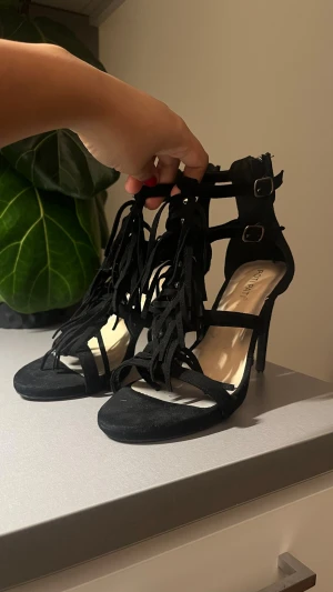 Svarta sandaler med fransar och klack - Säljer ett par svarta sandaler från Potipat med höga klackar och snygga fransdetaljer framtill. Skorna har öppen tå, spännen runt ankeln och är gjorda i mockaimitation. Perfekta för dig som vill sticka ut lite extra.
