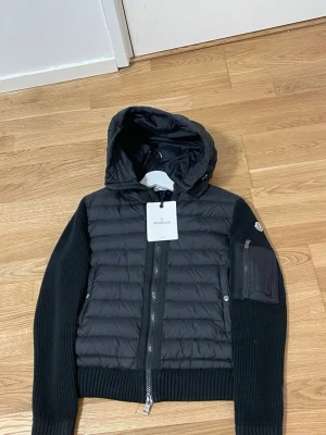 Svart pufferjacka  Moncler - Svart pufferjacka från Moncler med stickade ärmar och huva. Jackan har quiltad framsida, dragkedja, fickor med knappar och en extra ficka med dragkedja på ärmen. Materialmix av polyester och stickad ull ger en schysst kontrast. Perfekt för kyliga dagar.Liknande modell som orginala. 