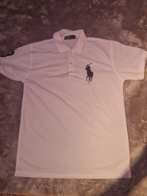 Sport pikétröja Polo Ralph Lauren M - Klassisk vit sport pikétröja från Polo Ralph Lauren i storlek M. Tröjan har korta ärmar, krage med tre knappar och den ikoniska stora polospelaren broderad på bröstet. Tillverkad i polyester och sitter tajt och bra.