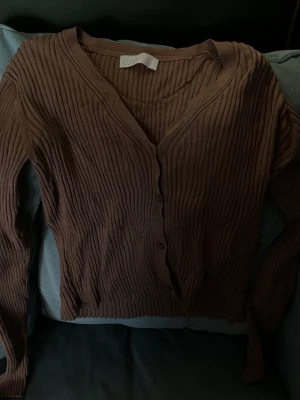 Ribbad brun kofta från Samsøe Samsøe - Säljer en ribbad kofta i brun nyans från Samsøe Samsøe. Koftan har v-ringning, knappar framtill och är långärmad. Materialet är mjukt och stretchigt, perfekt för lager på lager under höst och vår. Enkel och stilren design som passar till det mesta.