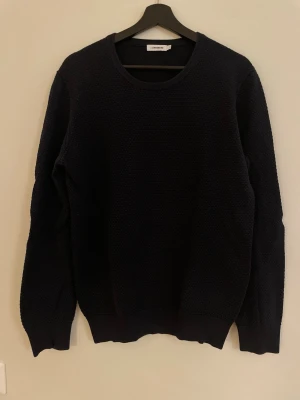 Mörkblå stickad tröja J.Lindeberg - Säljer en mörkblå stickad tröja från J.Lindeberg i storlek M. Tröjan har rund halsringning, diskret strukturmönster och är gjord i 100% bomull. Perfekt till höst och vinter för en clean och stilren look.