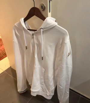 Vit hoodie från Moncler med half zip - Vit hoodie från Moncler i bomull med half zip och snörning i huvan. Långa ärmar, klassisk Moncler-logga på ärmen och coola rödblå ränder på insidan av huvan och vid ärmslut. Perfekt för dig som gillar stilrena och exklusiva plagg.