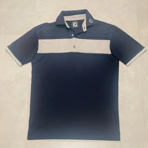 Footjoy golfpike  - Säljer en marinblå pikétröja från FootJoy med kontrasterande ljusgrå panel över bröstet och detaljer på kragen. Tröjan har korta ärmar, knappar framtill och klassisk krage. Perfekt för golf eller andra sportiga aktiviteter.