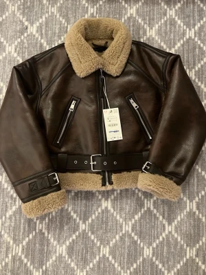 Brun teddyjacka med bälte och dragkedjor - Säljer en brun teddyjacka med beige foder och krage. Jackan har två snedställda dragkedjefickor, bälte i midjan och detaljer vid ärmslut. Utsidan är i skinnimitation och insidan är mjuk teddy. Perfekt för kalla dagar och riktigt snygg till jeans.