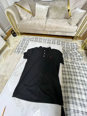 Svart piké från Raulph lauren - Säljer en svart pikétröja från Ralph Lauren med klassisk krage och tre vita knappar framtill. Tröjan har korta ärmar och den ikoniska röda logotypen broderad på bröstet. Perfekt till jeans eller shorts för en clean och stilren look.