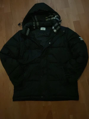 Svart pufferjacka från Burberry - Svart pufferjacka från Burberry med avtagbara ärmar, så du kan bära den som väst. Jackan har huva och klassiskt rutigt foder i halsen. Stora fickor framtill och snygga Burberry-detaljer på knappar och patch på ärmen. Perfekt för kalla dagar.