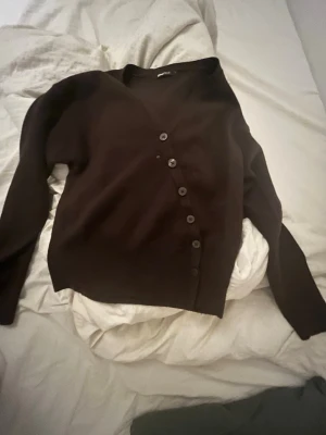 Brun stickad kofta med knappar - Mörkbrun stickad kofta med v-ringning och markerad midja. Koftan har långa, vida ärmar och stängs med en rad vita knappar framtill. Perfekt för lager på lager under kyligare dagar. Endast använd en gång