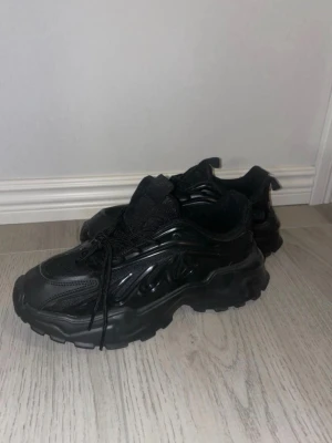 Chunky svarta sneakers med grov sula - Säljer ett par chunky svarta sneakers med riktigt grov och mönstrad sula. Skorna har snörning och en blandning av syntet och textil i ovandelen, vilket ger en cool och modern vibe. Perfekta för dig som gillar streetstyle och vill sticka ut lite extra.