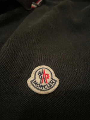 Svart Moncler piké - Svart pikétröja från Moncler med klassisk krage och knäppning framtill. Tröjan har Moncler-logga på bröstet och snygga rödvita detaljer på krage och ärmslut. Tillverkad i bomull för en skön känsla och stilren look.