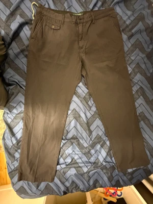 Bruna chinos från Henry Cotton's, stl 38 - Snygga bruna chinos från Henry Cotton's i storlek 38. Klassisk rak passform med knappdetalj på bakfickan och diskret logga. Tillverkade i mjuk bomull som känns skön mot huden. Perfekta för en clean och stilren look.