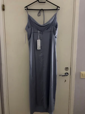 Ljusblå långklänning med tunna band - Säljer en elegant ljusblå långklänning med tunna axelband och snygg draperad urringning. Klänningen har en slät, glansig yta och knyt i nacken för extra detalj. Perfekt för dig som vill ha en stilren och modern look.