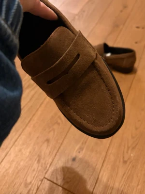 Bruna mockaloafers med rund tå - Snygga loafers i brun mocka från Zara med klassisk design och rund tå. Skorna har en platt sula och diskreta sömmar längs kanten. Perfekta att styla med jeans eller chinos för en chill och stilren look.