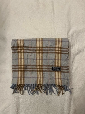 Burberry halsduk - Säljer en klassisk rutig halsduk från Burberrys i ljusblått, beige och brunt. Halsduken har någon litet litet små hål men d syns inte vid användning sjävklart för det knappt syns!