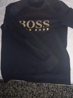 Svart sweatshirt från Hugo Boss - Svart sweatshirt från Hugo Boss med stort guldfärgat BOSS-tryck på bröstet. Tröjan har rund halsringning och långa ärmar. Materialet känns mjukt och bekvämt, perfekt för en avslappnad stil. Priset kan diskuteras 