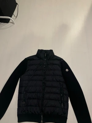 Moncler cardigan navy blue - Mörkblå cardigan från Moncler med stickade ärmar och quiltad front. Jackan har dragkedja framtill och två fickor med dragkedja. Moncler-logga på vänster ärm. Perfekt för dig som vill ha en snygg och stilren jacka med exklusiv känsla.