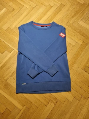 Blå sweatshirt från Huski Wear - Blå sweatshirt från Huski Wear med rund halsringning och ribbade muddar vid ärmslut och nederkant. Tröjan har en röd patch med vit text på vänster ärm och en liten diskret logga vid nederkanten. Perfekt för en avslappnad stil. Den är väldigt bekväm och mjuk att ha på sig.