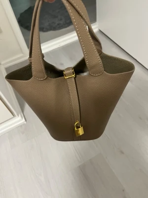 Beige handväska med gulddetaljer - Säljer en stilren beige/brun handväska i skinn med guldfärgat lås och spänne framtill. Väskan har två rejäla handtag och en mjukt strukturerad form som ger en exklusiv känsla. Aldrig använd!❤️