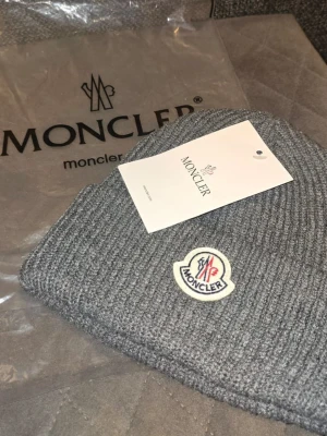 Grå ribbstickad mössa från Moncler - Snygg grå ribbstickad mössa från Moncler med klassisk Moncler-logga framtill. Mössan har en uppvikt kant och är tillverkad i mjukt material som håller dig varm. Perfekt accessoar för kyliga dagar och ger en stilren look. Priset är inte hugget i sten. Hör av er vid frågor. Helt ny mössa med påsen och tagen.