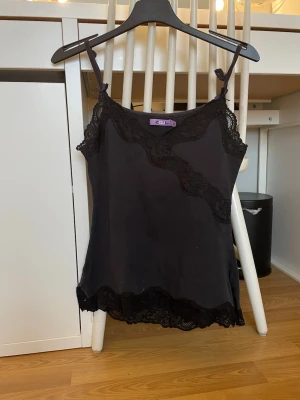 Svart spetslinnetopp från Zouli - Snygg svart linnetopp från Zouli med tunna axelband och dekorativa spetsdetaljer längs halsringning och nederkant. Toppen har en feminin v-ringad form och är perfekt för att styla lager på lager. Materialet känns mjukt och lätt.skulle säga passa xs/s är ganska liten i storlek💞