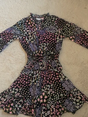 klänning från gina tricot  - Kort klänning med svart botten och blommigt mönster i rosa, lila, vitt och grönt. Klänningen har lång ärm, markerad midja med smock och lätt v-ringad hals. Materialet känns mjukt och stretchigt, troligen polyester eller bomull. Perfekt för vår och sommar.