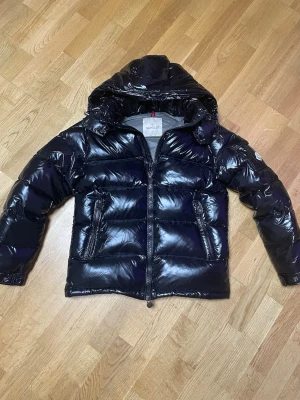 Moncler maya - Snygg svart dunjacka från Moncler med glansig finish och huva. Jackan har quiltad design, dragkedja framtill och två fickor med dragkedja. Perfekt för kalla dagar när du vill hålla stilen.