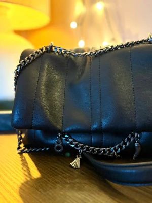 Svart quiltad handväska med kedja - Stilren svart handväska i quiltad design från Zara med silverfärgad kedja och dekorativa berlocker. Väskan har magnetlås och är tillverkad i mjukt skinnliknande material. Perfekt för dig som vill ha en trendig accessoar med coola detaljer.