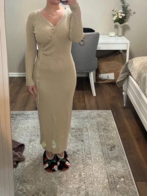 Beige ribbad omlottklänning - Säljer en beige långklänning med ribbad struktur och omlottdesign. Klänningen har lång ärm, v-ringning och snörningsdetalj i sidan för en snygg passform. Perfekt för dig som gillar stilrena och bekväma plagg.