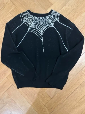 Stockholm surfboard club  - Cool svart stickad tröja med vitt spindelnätsmönster över axlar och bröst. Tröjan har rund halsringning och långa ärmar. Perfekt för dig som gillar unika detaljer och vill sticka ut lite extra. Två små hål från slitage längst ner på armen, syns knappt.