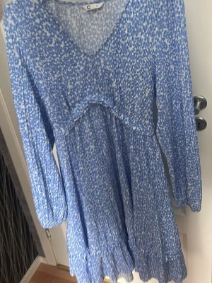 Blåvit blommig klänning med v-ringning - Söt blåvit klänning med blommigt mönster och v-ringad hals. Klänningen har lång ärm och lätt rynkad kjol för en flowy känsla. Perfekt för vår och sommar, tillverkad i ett mjukt och luftigt material.