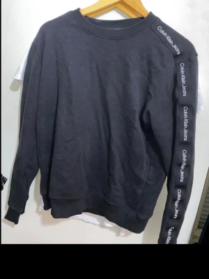 Svart sweatshirt från Calvin Klein Jeans - Svart sweatshirt med rund hals och ribbade muddar från Calvin Klein Jeans. Längs ena ärmen finns en vit logga tryckt upprepade gånger. Tröjan har en avslappnad passform och är tillverkad i mjukt bomullsmaterial. Perfekt för en casual och trendig look.