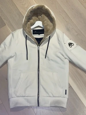 Vit dunjacka från Moose Knuckles - Snygg vit dunjacka från Moose Knuckles med huva fodrad i beige fuskpäls och svarta snören. Jackan har dragkedja framtill, två fickor och en ikonisk logga på ärmen. Insidan är quiltad och svart med diskret mönster. Perfekt för kalla dagar.