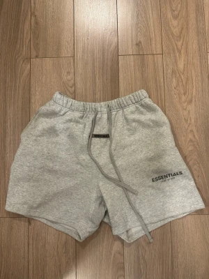 Grå Essentials shorts med snörning - Mysiga grå shorts från Essentials med elastisk midja och snörning framtill. Klassisk passform med två sidofickor och en bakficka. Essentials-logga tryckt på vänster ben. Perfekta för chill eller träning. Shortsen är lite noppriga