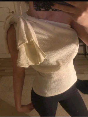 Vit oneshoulder topp med volang - Säljer en stilren vit oneshoulder topp med volangdetalj på axeln. Toppen är ribbad och har en tight passform som framhäver figuren. Perfekt för dig som vill ha något unikt och trendigt i garderoben.