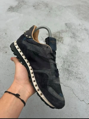 Valentino Garavani  - Säljer ett par svarta och mörkgrå Valentino Garavani Rockrunner sneakers med coolt kamouflagemönster och ikoniska nitar runt sulan. En nit saknas på baksidan! ‼️ Skorna har ovandel i mocka och textil, svarta skosnören och en chunky gummisula. Perfekta för dig som vill sticka ut med stil.